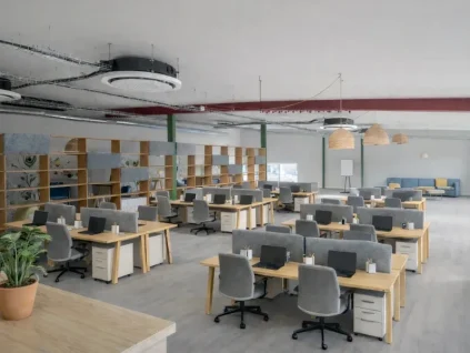 Espace de coworking en open space aménagé avec des rangées de bureaux en bois, des séparateurs acoustiques gris et une grande étagère de séparation, sous un plafond de style industriel.