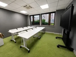 Intérieur de la salle Bagdad au BURO Club Partner Dijon Centre, une salle de réunion ou formation avec moquette verte, longues tables blanches pliables et chaises blanches, murs gris et grand écran interactif sur pied roulant.