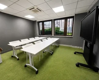 Intérieur de la salle Bagdad au BURO Club Partner Dijon Centre, une salle de réunion ou formation avec moquette verte, longues tables blanches pliables et chaises blanches, murs gris et grand écran interactif sur pied roulant.