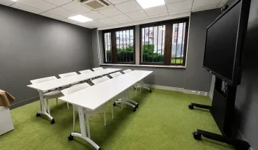 Intérieur de la salle Bagdad au BURO Club Partner Dijon Centre, une salle de réunion ou formation avec moquette verte, longues tables blanches pliables et chaises blanches, murs gris et grand écran interactif sur pied roulant.