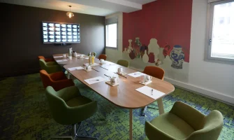 Ambiance colorée dans La Loge Tigre et Dragon du BURO Club Partner Dijon Centre, une salle de réunion avec moquette verte, chaises en velours vert et orange, table ovale en bois et fresque murale représentant des vases asiatiques sur un mur rouge vif.
