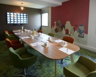 Ambiance colorée dans La Loge Tigre et Dragon du BURO Club Partner Dijon Centre, une salle de réunion avec moquette verte, chaises en velours vert et orange, table ovale en bois et fresque murale représentant des vases asiatiques sur un mur rouge vif.