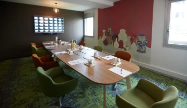 Ambiance colorée dans La Loge Tigre et Dragon du BURO Club Partner Dijon Centre, une salle de réunion avec moquette verte, chaises en velours vert et orange, table ovale en bois et fresque murale représentant des vases asiatiques sur un mur rouge vif.
