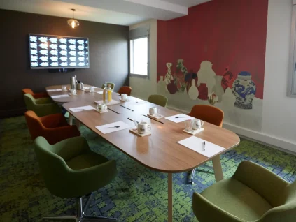Ambiance colorée dans La Loge Tigre et Dragon du BURO Club Partner Dijon Centre, une salle de réunion avec moquette verte, chaises en velours vert et orange, table ovale en bois et fresque murale représentant des vases asiatiques sur un mur rouge vif.