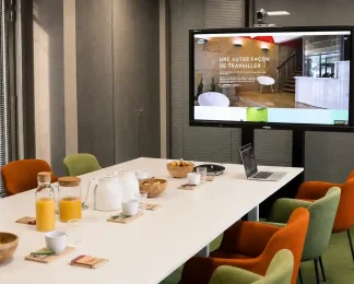 Zoom sur la table de réunion de la salle Palmyre au BURO Club Partner Dijon Centre, équipée pour la visioconférence avec chaises en velours, grand écran Vivitek affichant "Une autre façon de travailler", et collations.