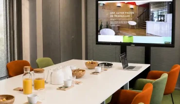 Zoom sur la table de réunion de la salle Palmyre au BURO Club Partner Dijon Centre, équipée pour la visioconférence avec chaises en velours, grand écran Vivitek affichant "Une autre façon de travailler", et collations.
