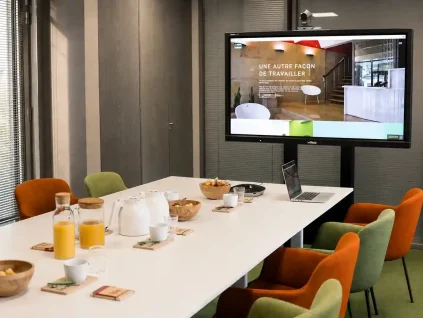 Zoom sur la table de réunion de la salle Palmyre au BURO Club Partner Dijon Centre, équipée pour la visioconférence avec chaises en velours, grand écran Vivitek affichant "Une autre façon de travailler", et collations.