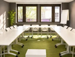 Vue de la salle Petra au BURO Club Partner Dijon Centre, une salle de réunion en U lumineuse avec moquette verte, tables blanches, chaises pliables blanches et grandes fenêtres donnant sur la ville ensoleillée.