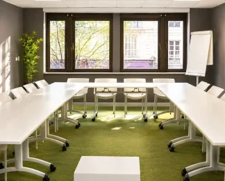 Vue de la salle Petra au BURO Club Partner Dijon Centre, une salle de réunion en U lumineuse avec moquette verte, tables blanches, chaises pliables blanches et grandes fenêtres donnant sur la ville ensoleillée.