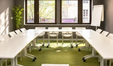Vue de la salle Petra au BURO Club Partner Dijon Centre, une salle de réunion en U lumineuse avec moquette verte, tables blanches, chaises pliables blanches et grandes fenêtres donnant sur la ville ensoleillée.