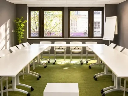 Vue de la salle Petra au BURO Club Partner Dijon Centre, une salle de réunion en U lumineuse avec moquette verte, tables blanches, chaises pliables blanches et grandes fenêtres donnant sur la ville ensoleillée.