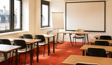 Vue de la salle Zanzibar au BURO Club Partner Dijon Centre, une grande salle de formation lumineuse avec moquette rouge, rangées de tables individuelles en bois et chaises noires, paperboard et grand écran de projection au fond.